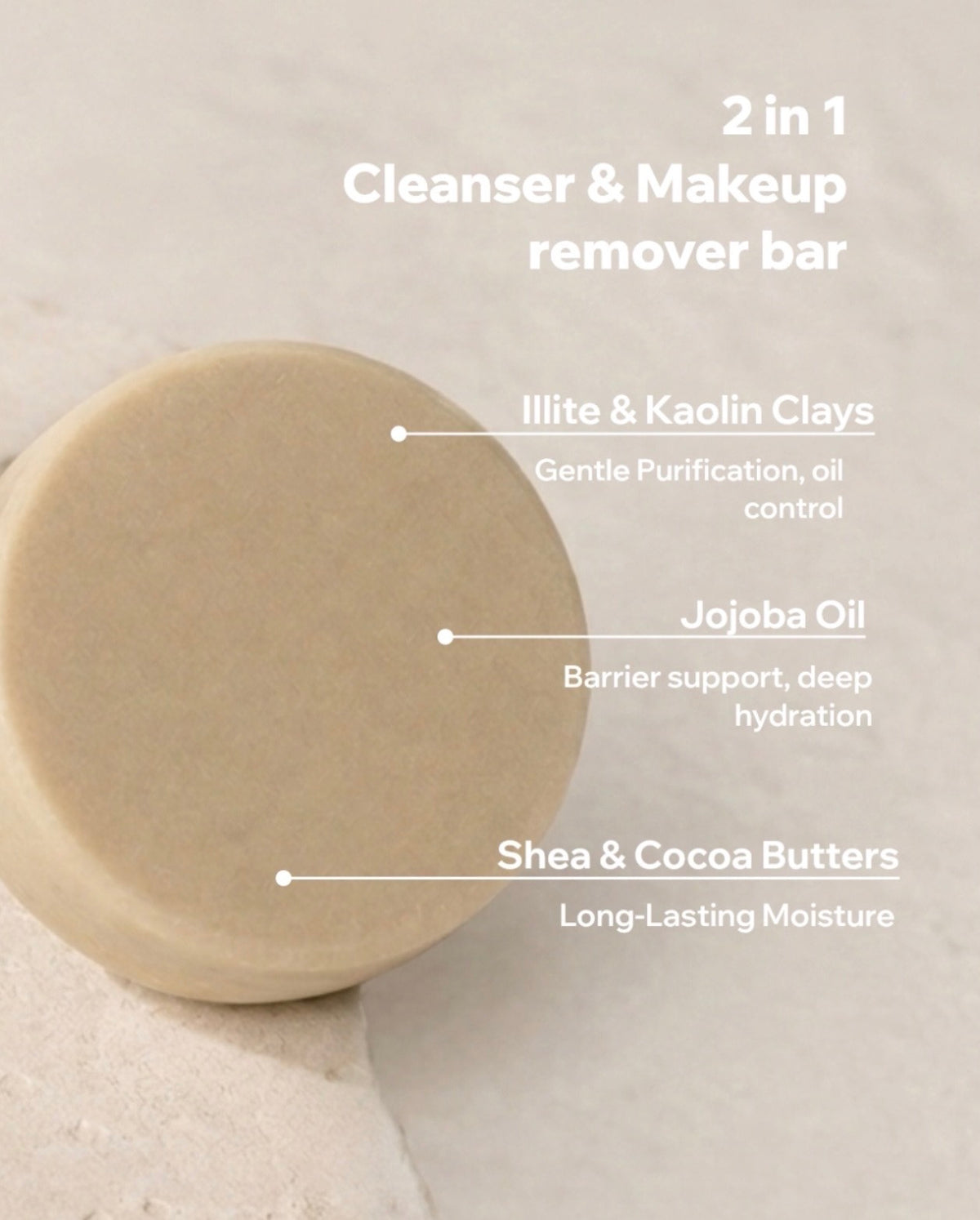 A round beige cleanser and makeup remover bar on a light textured surface with white text overlays describing its ingredients: Illite & Kaolin Clays, Jojoba Oil, and Shea & Cocoa Butters.