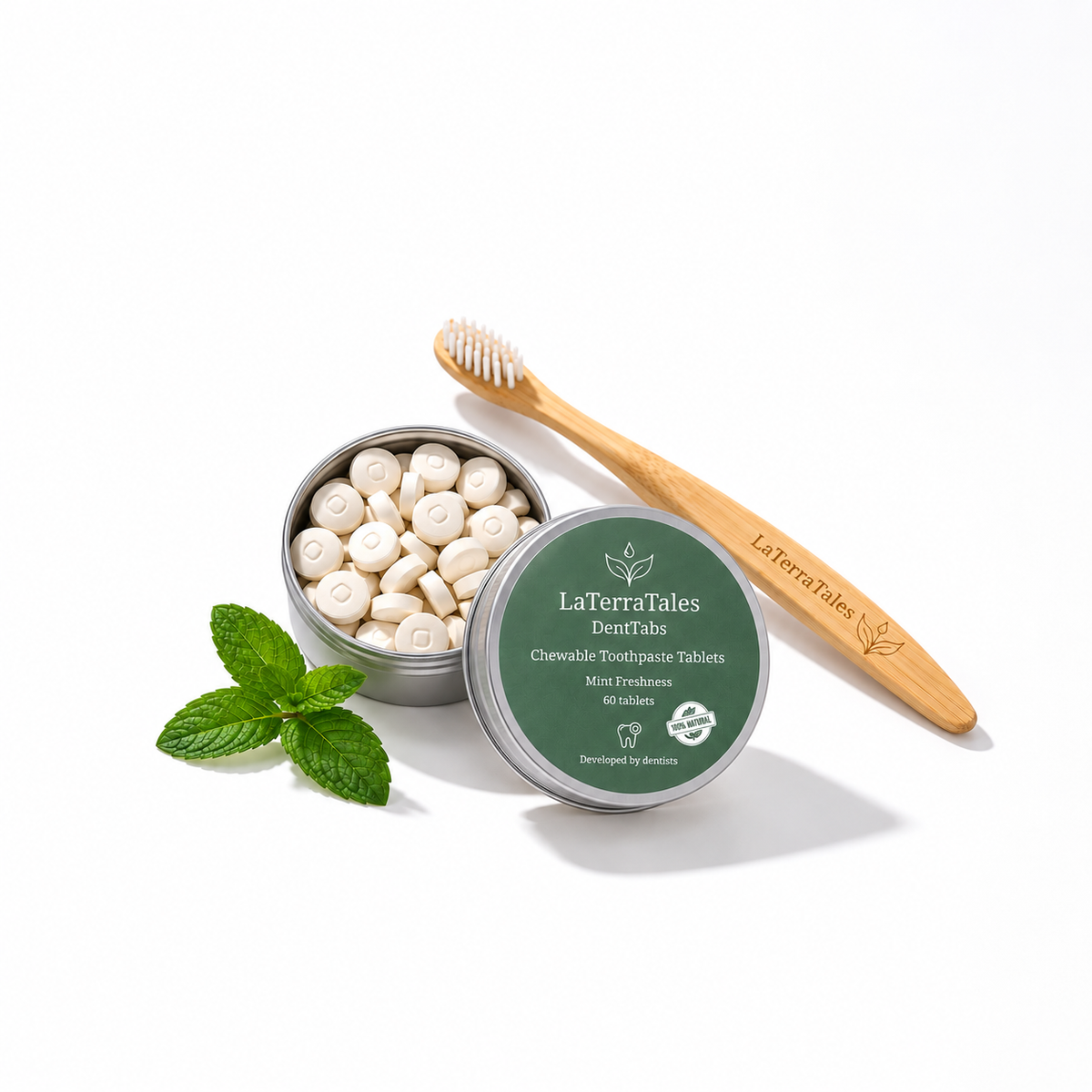 LaTerraTales DentTabschewable toothpaste tablets in an open silver tin, shown with a bamboo toothbrush and mint leaves on a white background.
