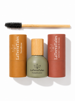 LaTerraTales hair and beauty set on a white background, featuring a black-and-bamboo spoolie brush, a terracotta brown tube labeled TerraBrow, a sage green bottle labeled Tranquility Elixir with a bamboo cap, and a terracotta orange tube labeled Amice Lip & Cheeks.